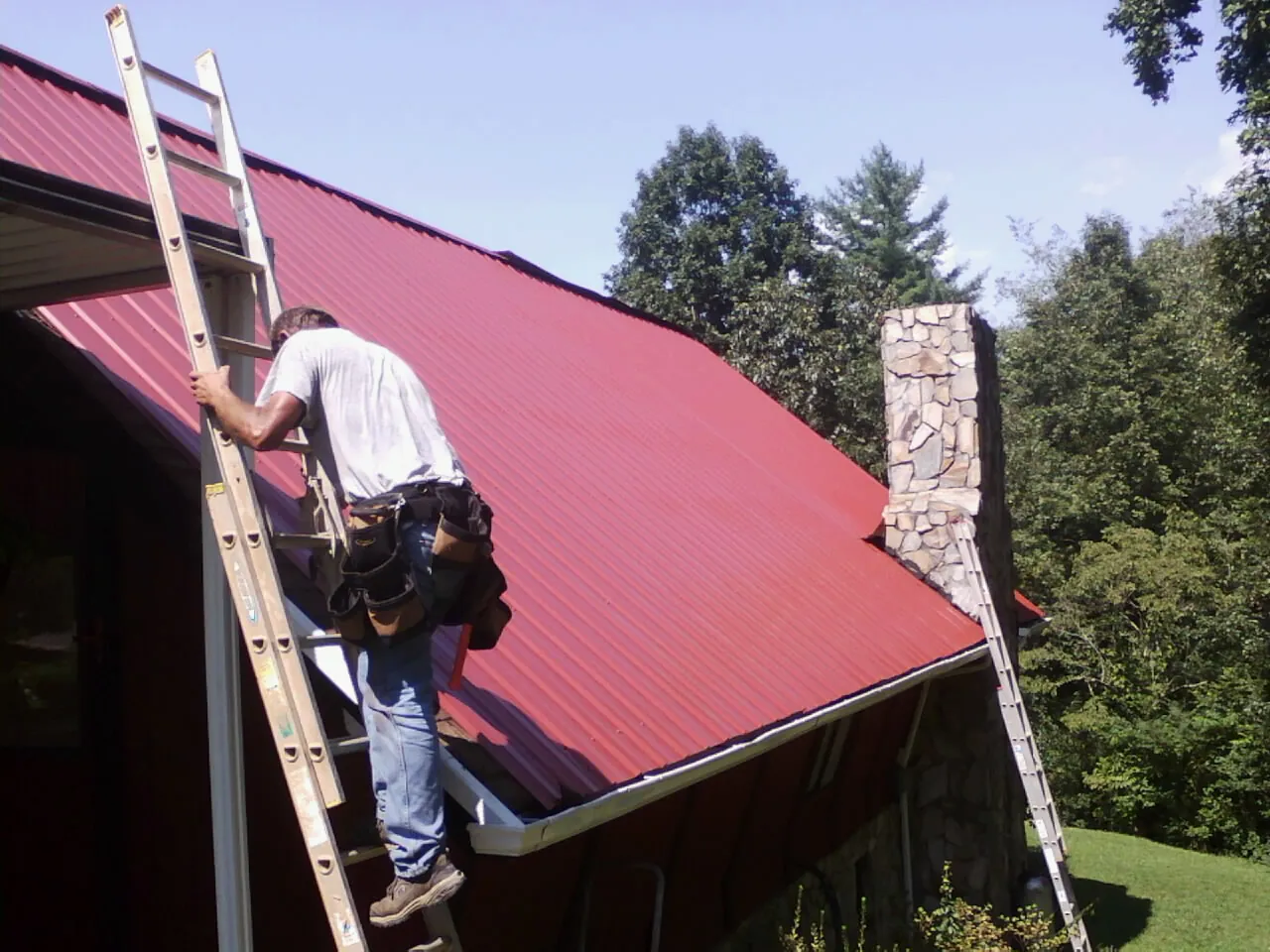 Roofer on a ladder beside a red metal roof and stone chimney — Soffit & Fascia Repair by Roofing Brothers in Poncha Springs